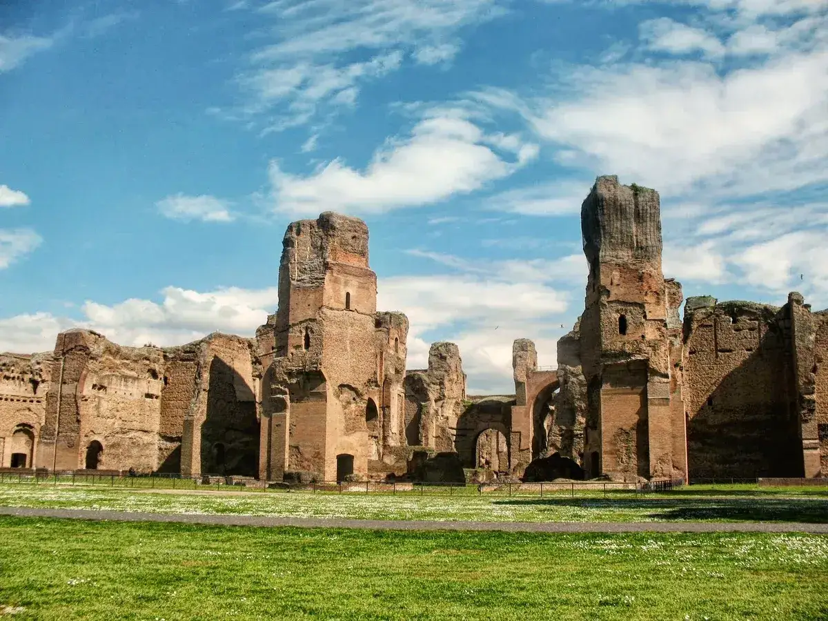 Ruines imposantes du **bain de Caracalla** sous un ciel bleu et nuageux. L'herbe verte et les petites fleurs bordent les vestiges antiques.