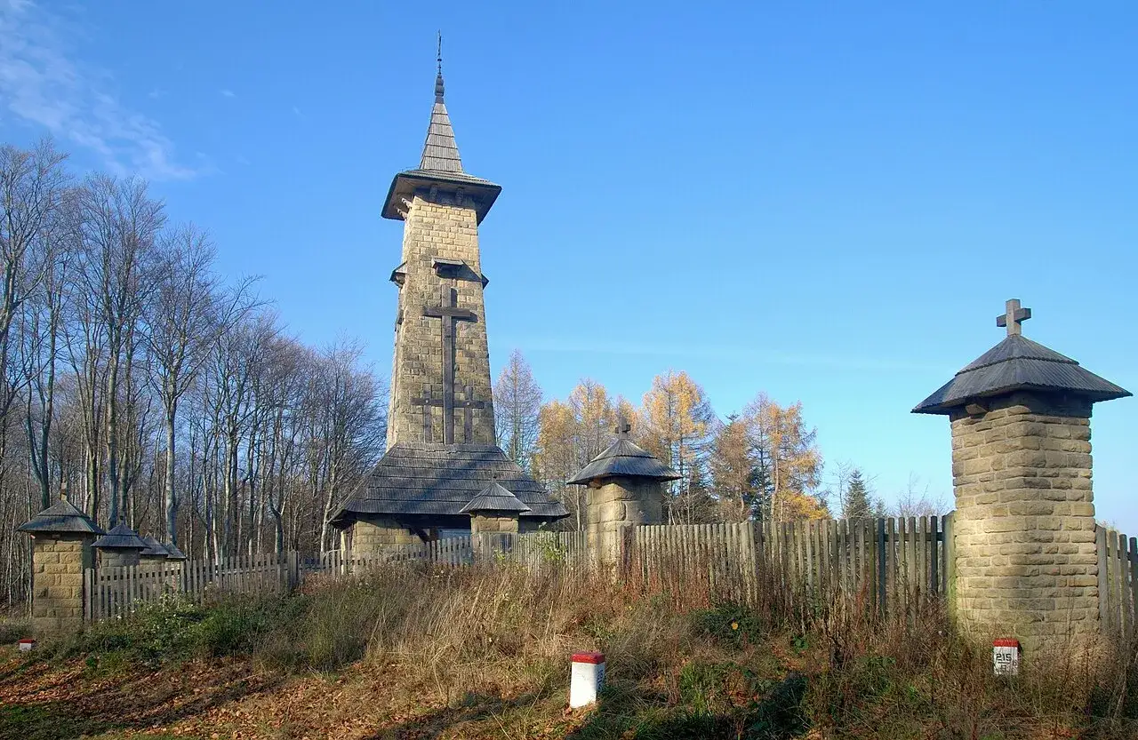 Kamienna wieża z krzyżem i drewniane kapliczki, otoczone płotem i drzewami. To jedne z **regietów atrakcje** w jesiennym krajobrazie.