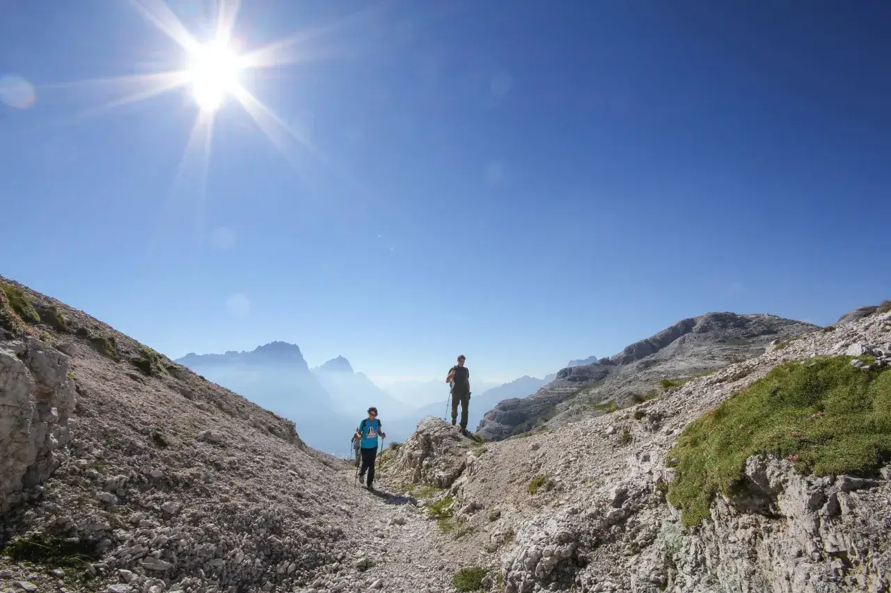 Due escursionisti percorrono il sentiero dei camosci sotto un sole splendente, con montagne nebbiose all'orizzonte.
