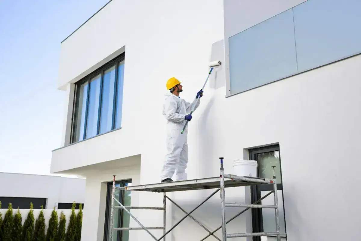 Hombre con casco amarillo y traje blanco realiza pintura mural exterior en una casa moderna, subido a un andamio.