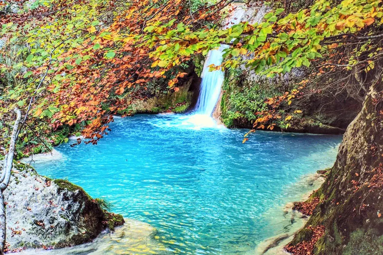 Cascada cayendo en una **piscina natural** de agua turquesa, rodeada de &aacute;rboles con hojas oto&ntilde;ales.