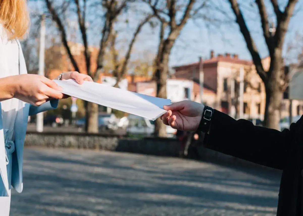 Dwie osoby przekazują sobie białą ulotkę informacyjną na tle drzew i budynków.