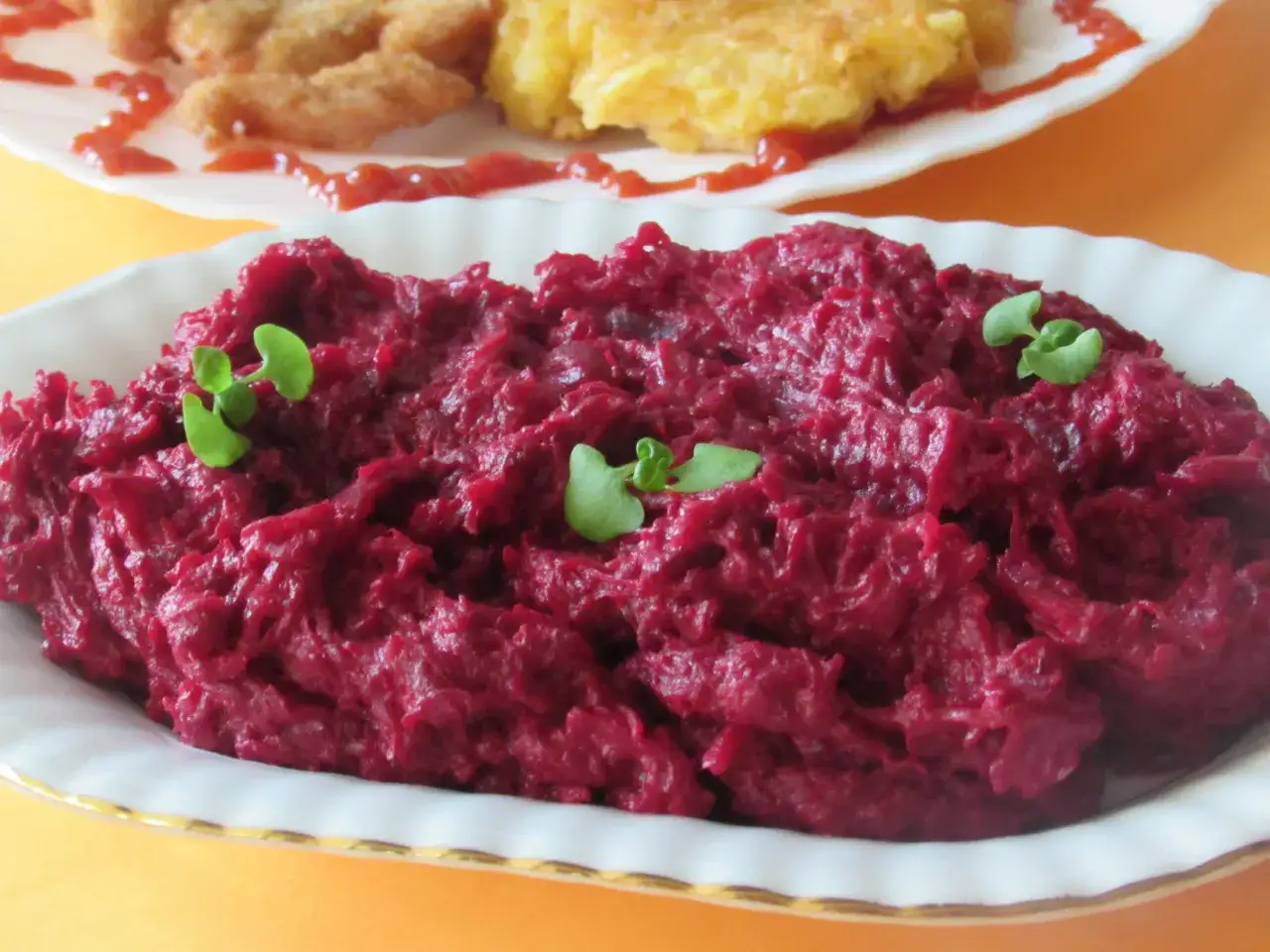 Pyszne, purpurowe buraczki na ciepło, udekorowane listkami ziół. W tle widać kotlety i placki ziemniaczane.