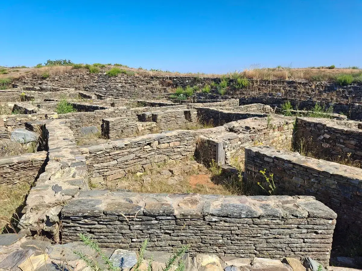 Ruinas de piedra del castro de Castromaior bajo un cielo azul. Muros bajos y senderos de grava se extienden por el yacimiento arqueol&oacute;gico.