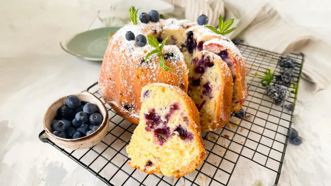 Saftiger Joghurt Kuchen mit Blaubeeren, bestäubt mit Puderzucker und garniert mit frischer Minze. Ein Stück ist bereits angeschnitten.
