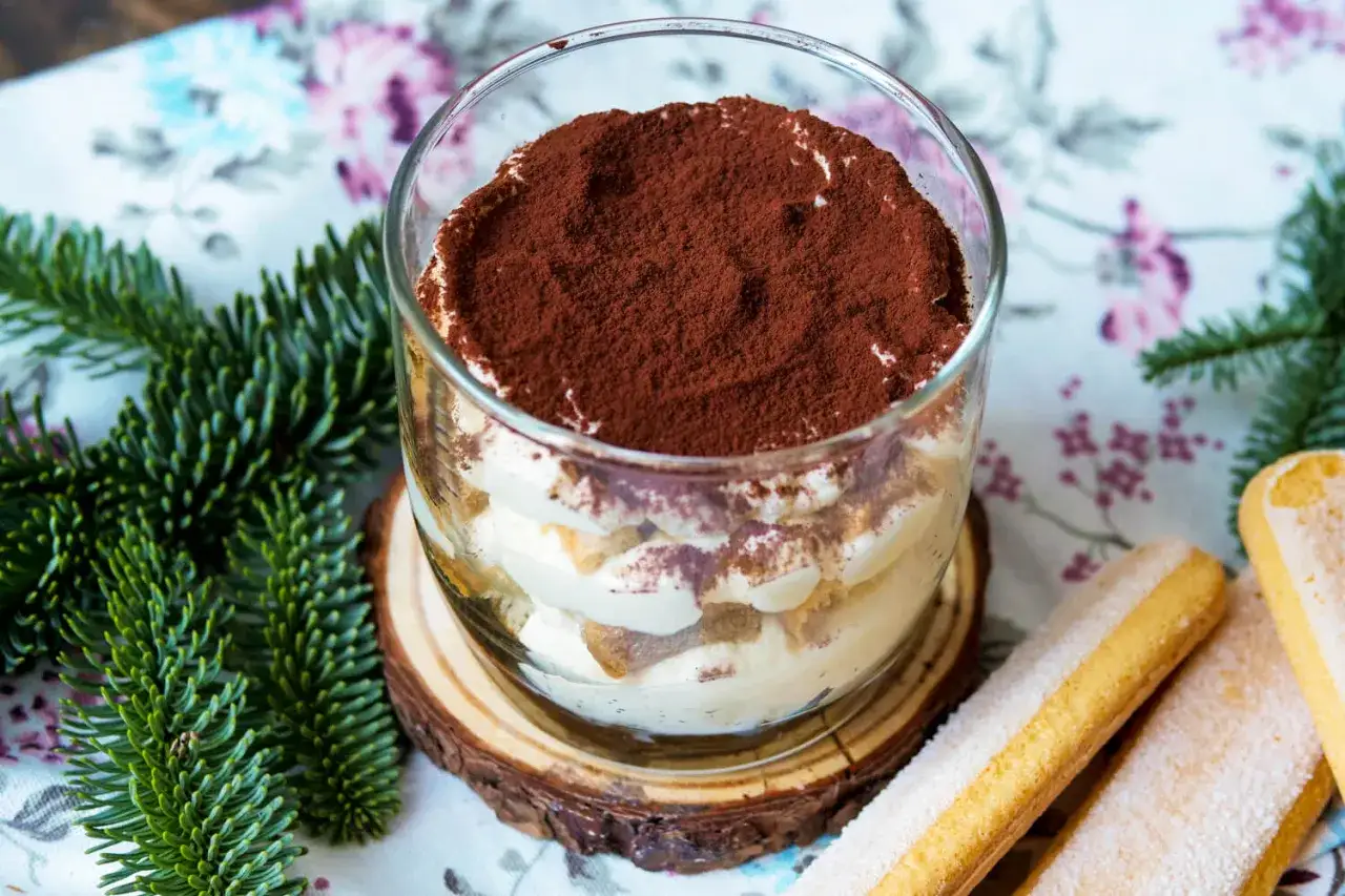 Cremiges Tiramisu ohne Ei im Glas, best&auml;ubt mit Kakao. Daneben liegen L&ouml;ffelbiskuits und Tannenzweige.