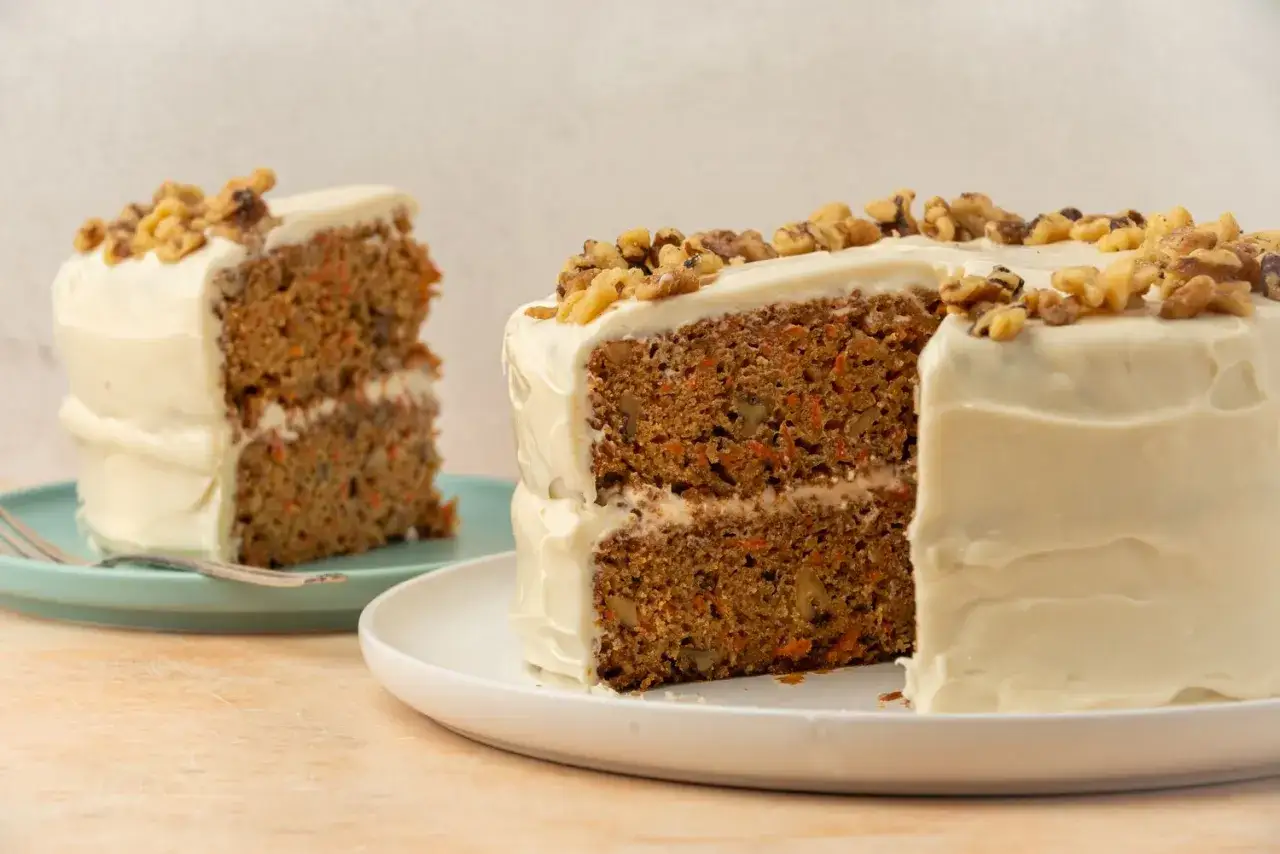 En saftig vegansk morotskaka med krämig frosting och valnötter. En bit är uppskuren och visas på en blå tallrik.