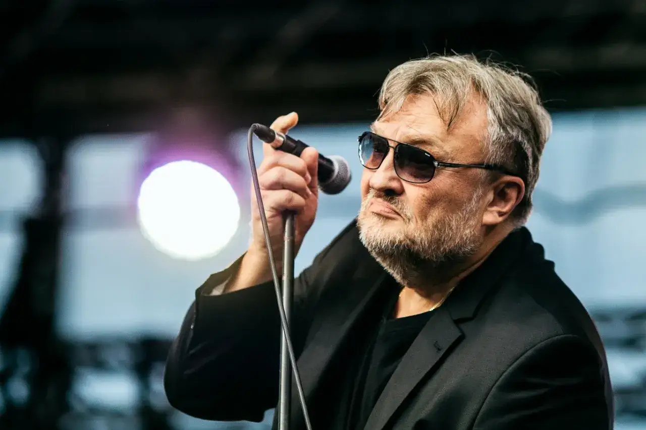 Wokalista Budki Suflera, Krzysztof Cugowski, w okularach przeciwsłonecznych śpiewa do mikrofonu na koncercie.