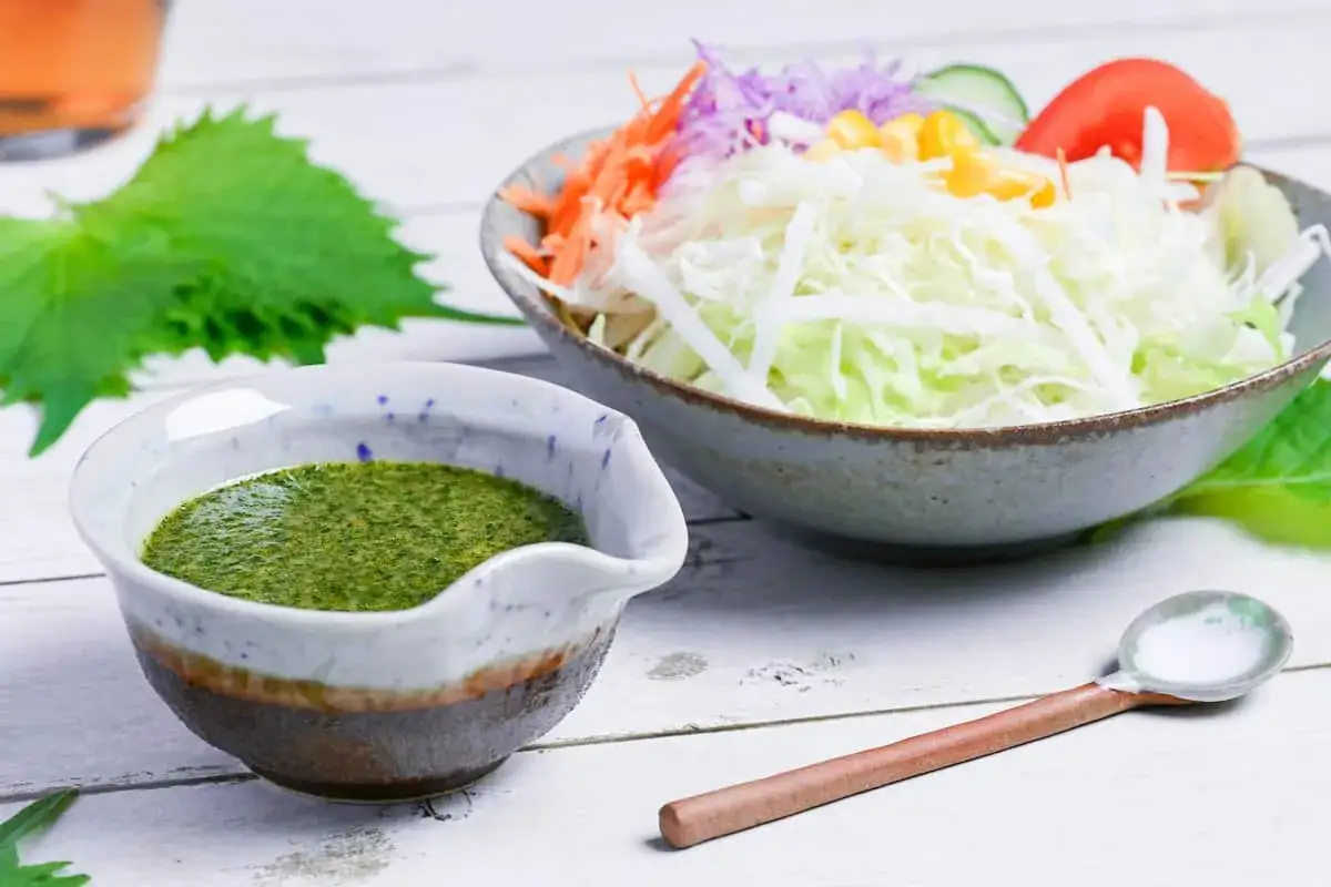 A vibrant green wasabi dressing sits in a ceramic bowl next to a fresh salad. A wooden spoon rests nearby, ready to serve.