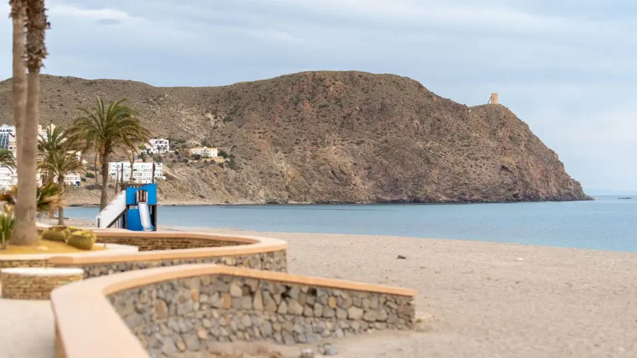 La playa El Ancón en Carboneras, con su arena dorada, palmeras y un tobogán azul, se extiende hacia un mar sereno y una montaña coronada por una torre.