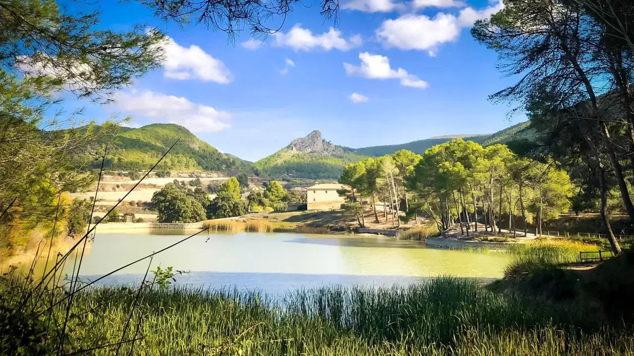 El bosquet de Moixent, un lago sereno rodeado de pinos y montañas verdes, con una casa y un pico rocoso al fondo.