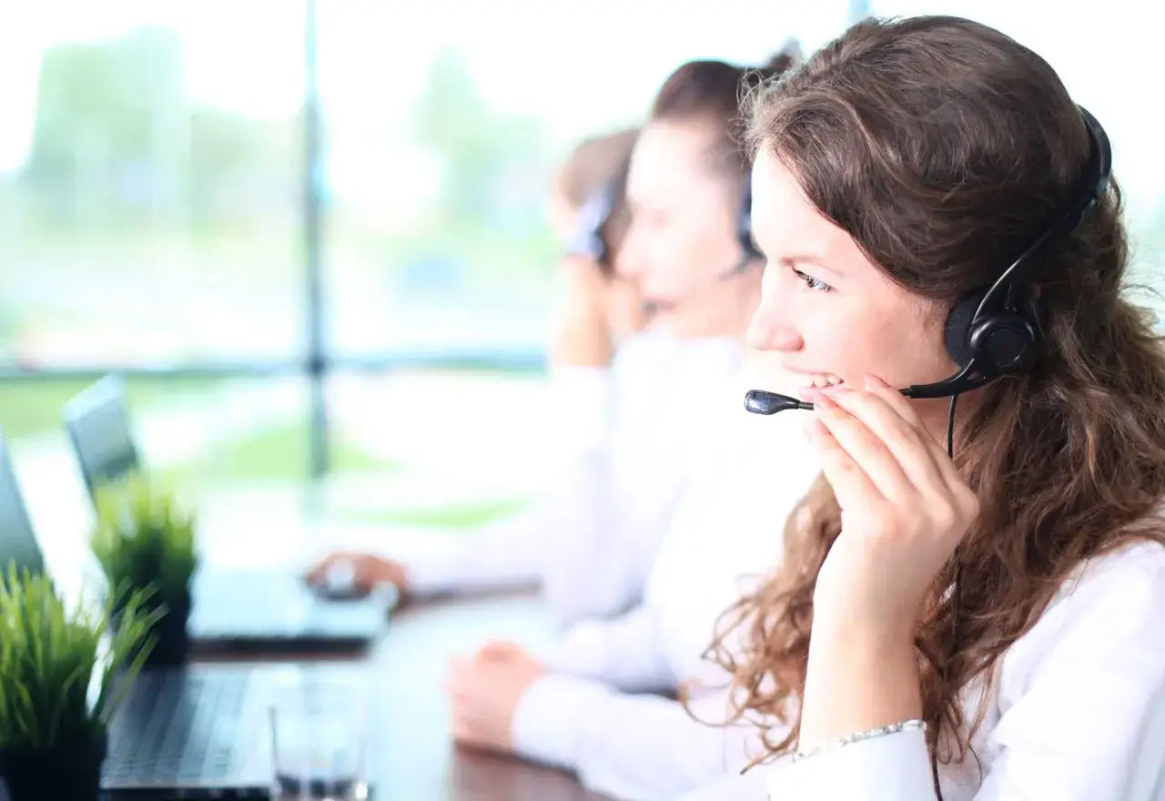 Callcenter-Mitarbeiterin mit Headset, die lächelnd spricht. Im Hintergrund Kolleginnen und Laptops. Kundenservice ist hier mos wert.