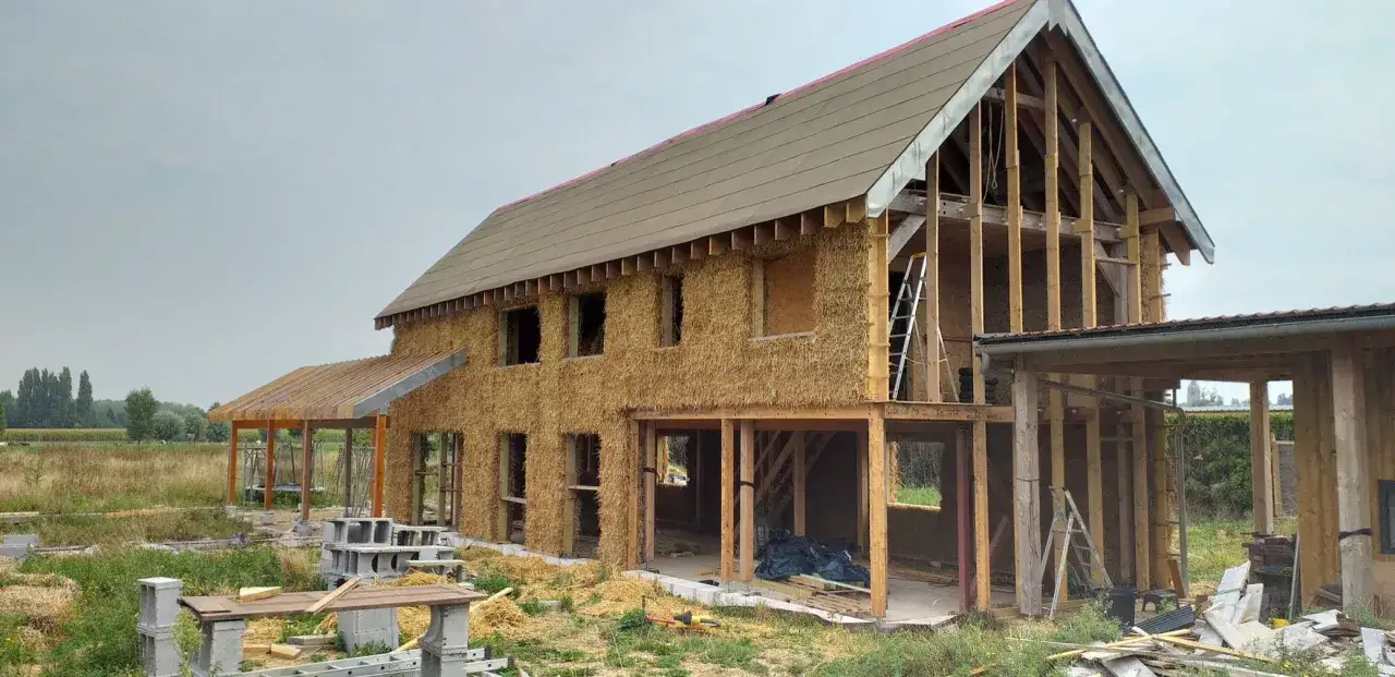 Construction d'une maison en paille, avec murs isolés et charpente visible. Le chantier est en cours, entouré de matériaux de construction.