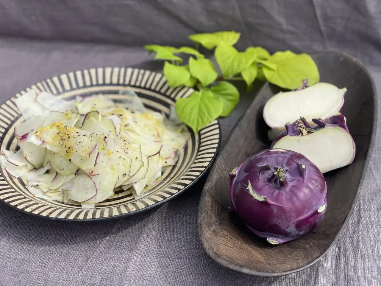En skål med kålrabbi-sallad, en hel lila kålrabbi och en delad kålrabbi i en träskål. Gröna blad i bakgrunden. Ett enkelt kålrabbi recept.
