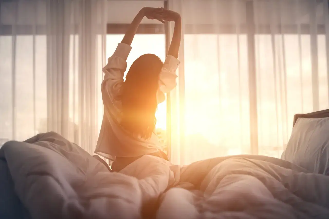 A woman stretches in bed as the sun rises, a peaceful scene that makes you wonder how long does it take to catch up on sleep.