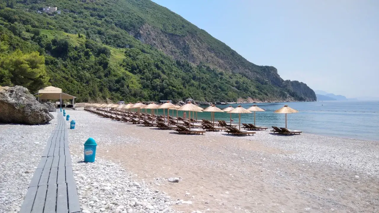 Plaża Jaz wita rzędem leżaków i parasoli. W tle zielone wzgórza i spokojne morze. Idealne miejsce na relaks.