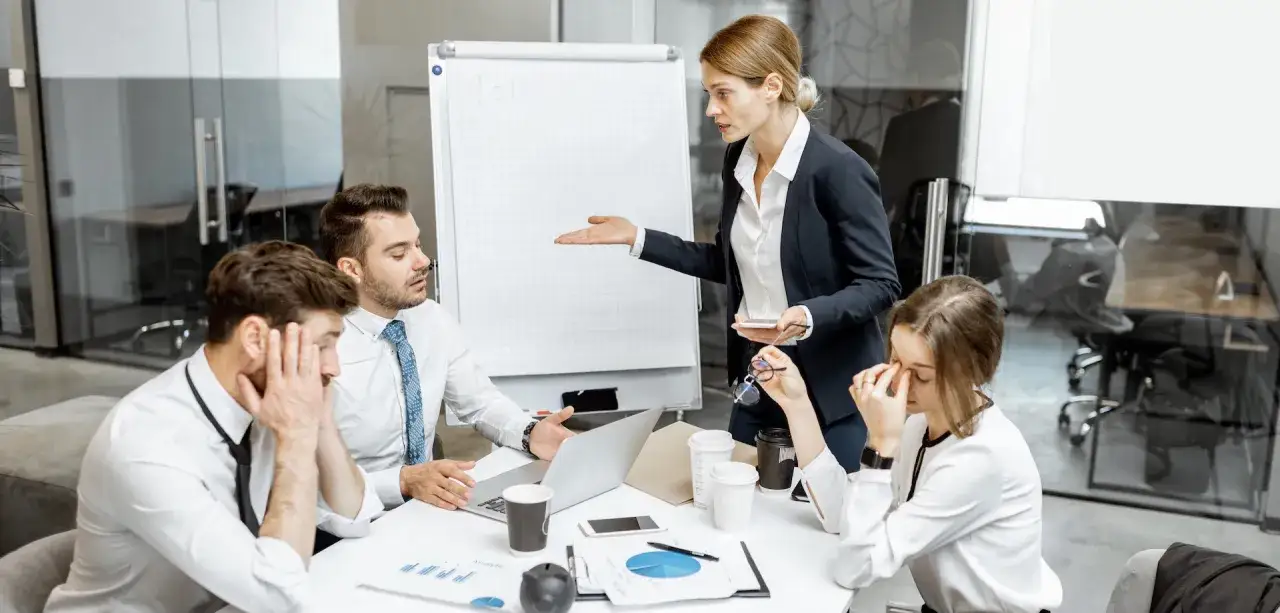 En kvinna gestikulerar mot en whiteboard medan kollegor ser utmattade ut. En scen som illustrerar passiv aggressiv kommunikation p&aring; arbetsplatsen.