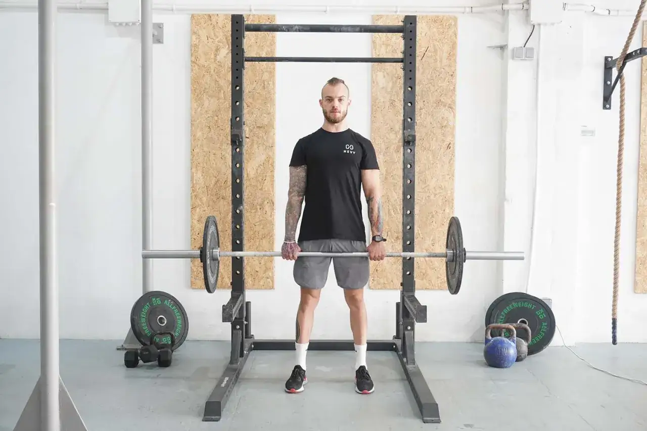 Mann mit Bart und Tattoos steht vor einem Power Rack, bereit f&uuml;r Rack Pulls. Hantelscheiben und Kettlebells liegen daneben.