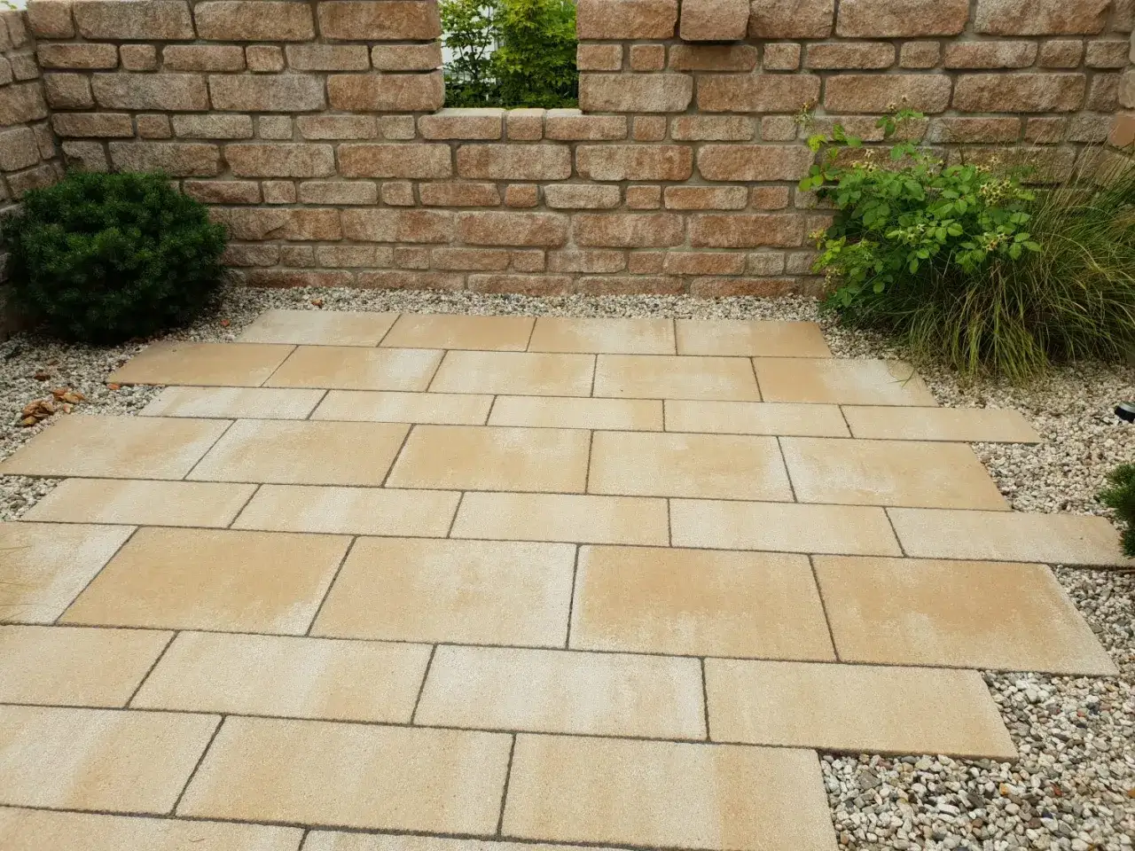 Eine Terrasse aus hellen Steinplatten und Kies, flankiert von einer Ziegelmauer und gr&uuml;ner Bepflanzung. Hier sieht man, was ist Trass: eine sch&ouml;ne Gestaltung f&uuml;r Au&szlig;enbereiche.
