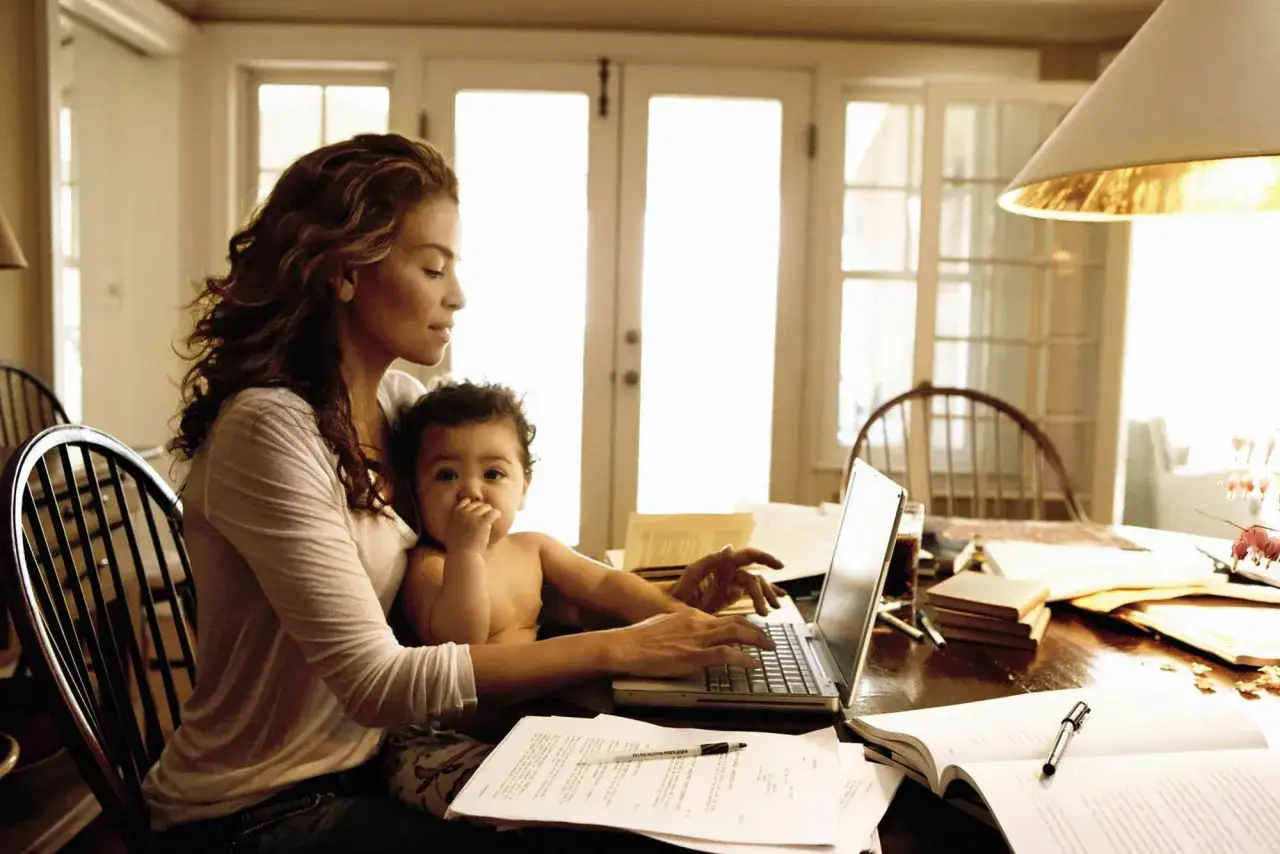 Una mamma lavoratrice con il suo bambino in grembo, mentre digita al computer tra documenti e libri.