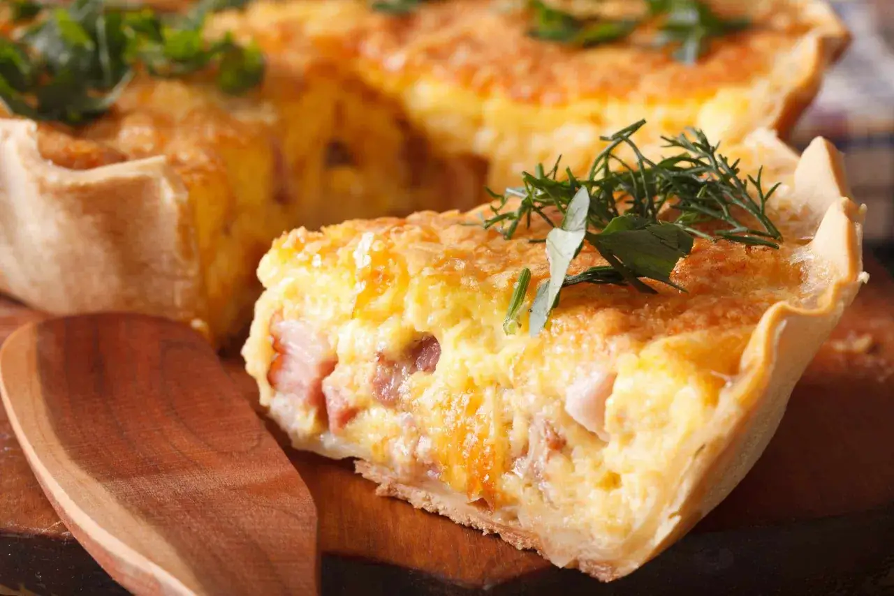 Une part de tarte raclette sans pomme de terre, garnie de lardons et d'herbes fraîches, sur une planche en bois.