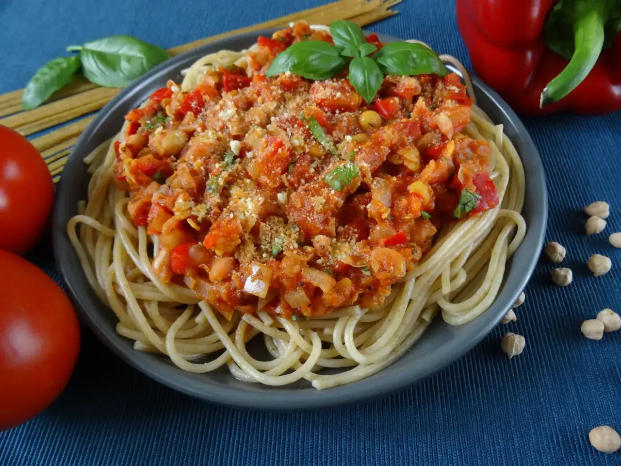 Makaron z ciecierzycy w sosie pomidorowym z papryką i bazylią, posypany parmezanem. Obok leżą pomidory i ciecierzyca.