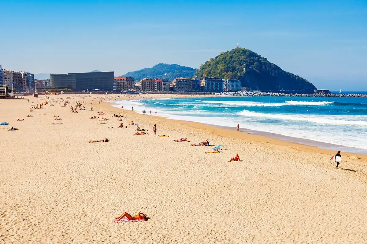 Słoneczna plaża w San Sebastian, z ludźmi odpoczywającymi na piasku i surferami na falach. W tle widać zielone wzgórze z pomnikiem.