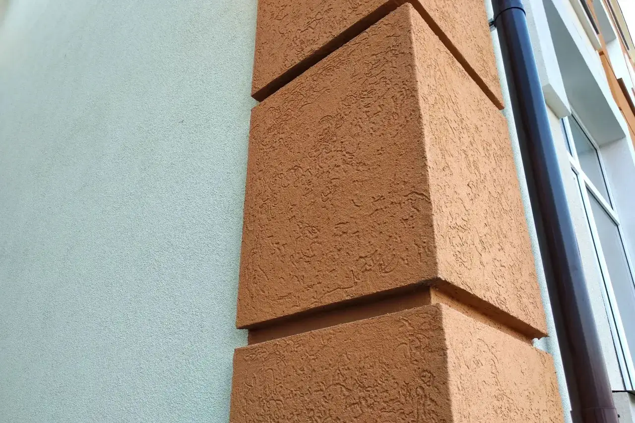 Brązowy tynk mozaikowy na narożniku budynku, obok jasnoszarej ściany. Widać rynnę i fragment okna.