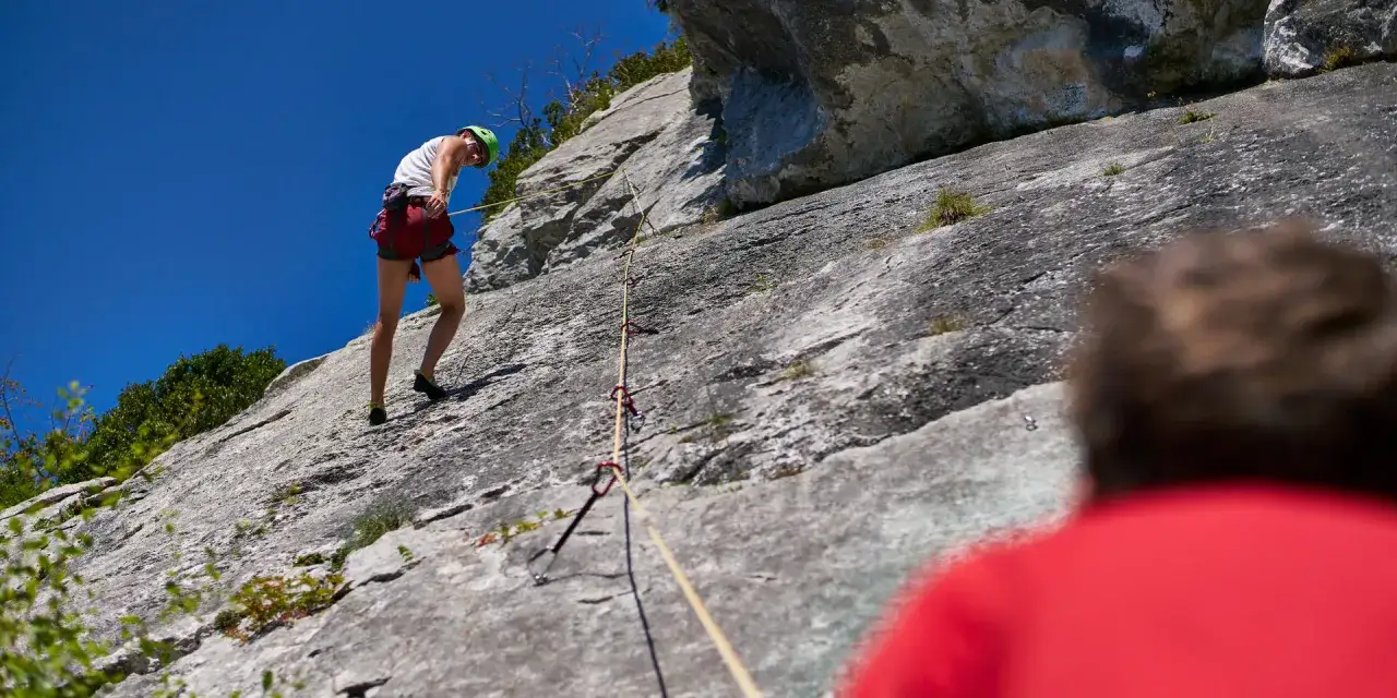 Un grimpeur en plein effort d'escalade 64, harnaché et casqué, progresse sur une paroi rocheuse sous un ciel bleu éclatant.