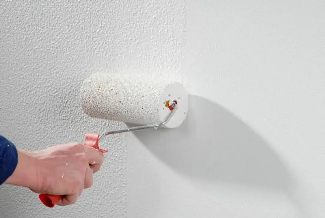Eine Hand mit Farbroller trägt eine weiße Grundierung auf eine strukturierte Wand auf. Rollputz Grundierung notwendig für ein gleichmäßiges Ergebnis.