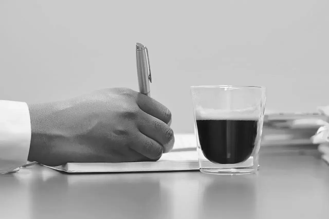 Dłoń pisze długopisem na papierze, obok szklanka kawy. Może to być testament.