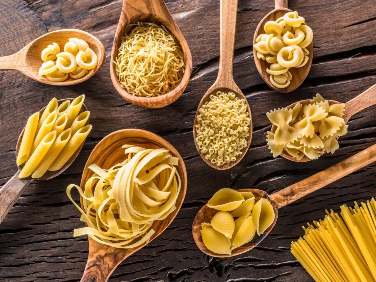 Drewniane łyżki pełne różnych rodzajów makaronu: penne, farfalle, muszelki, spaghetti, tagliatelle i drobne makarony.