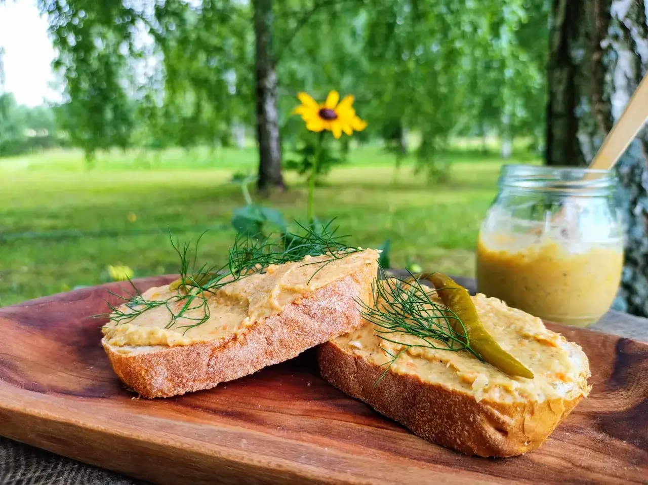 Dwa kawałki chleba z pasztetową, udekorowane koperkiem i ogórkiem. Słoik z pasztetową w tle. Idealna przekąska na piknik.