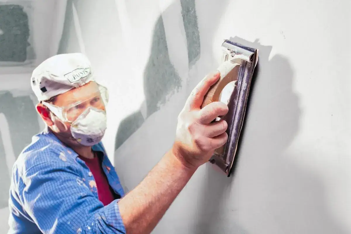 Hombre con mascarilla y gafas protectoras, lijando yeso en una pared para dejarla lisa.