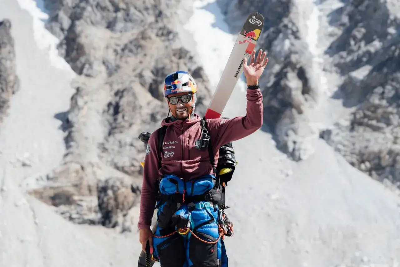 Kacper Tekieli, mit Skiern auf dem Rücken und winkend, steht vor einer schneebedeckten Bergkulisse.