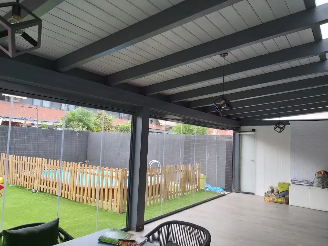 Un porche acristalado con vistas a un jardín con piscina infantil y césped artificial. El techo de madera y vigas oscuras, junto a lámparas modernas, crean un ambiente acogedor.