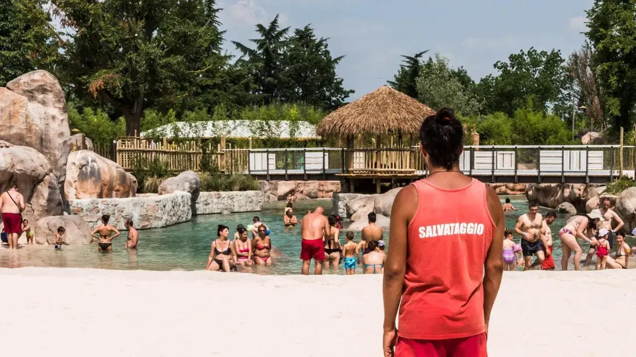Bagnino in divisa rossa sorveglia la piscina con bagnanti. Chiss&agrave; quanto guadagna un bagnino per questo lavoro estivo.