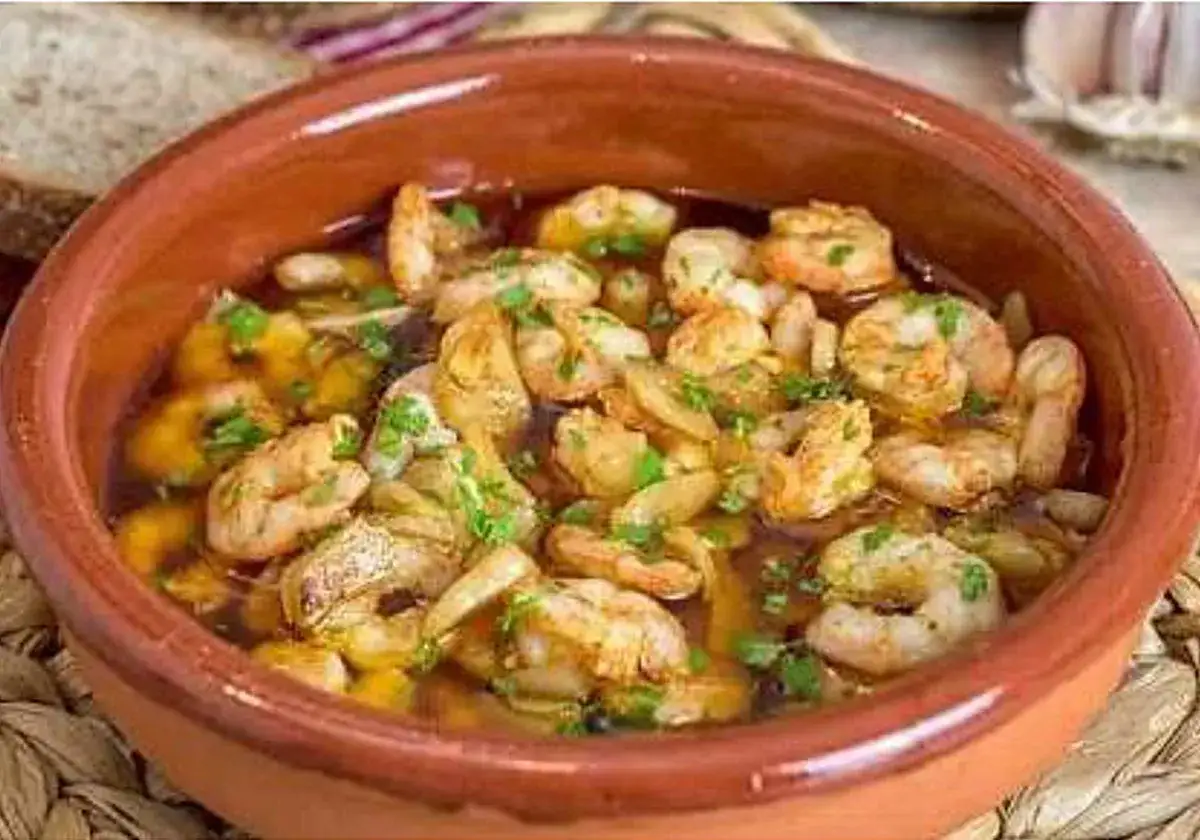 Un cuenco de barro rebosante de gambas al ajillo, con trozos de pan y un ajo al fondo.