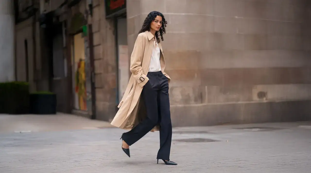Donna con trench beige e pantalone nero elegante, cammina per strada. Il pantalone significato è eleganza e praticità.