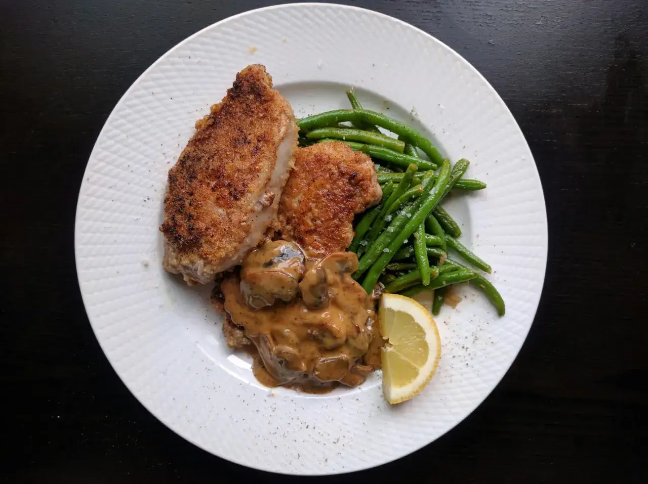 En panerad kotlett serveras med krämig svampsås, gröna bönor och en citronklyfta.