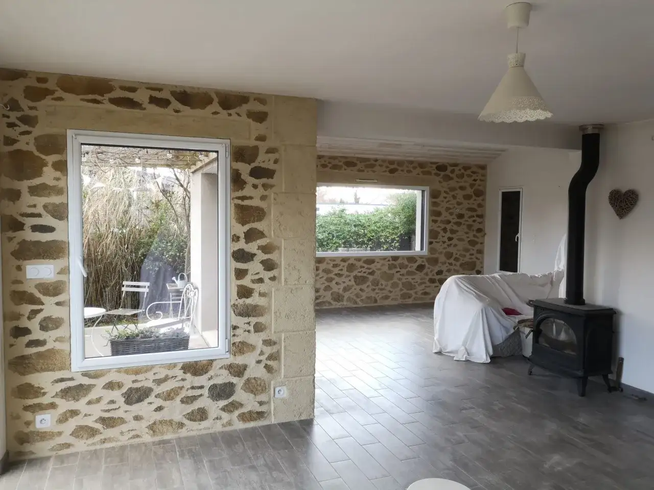 Un salon lumineux avec un mur en pierre apparente, un poêle à bois noir et un canapé recouvert d'un drap blanc.