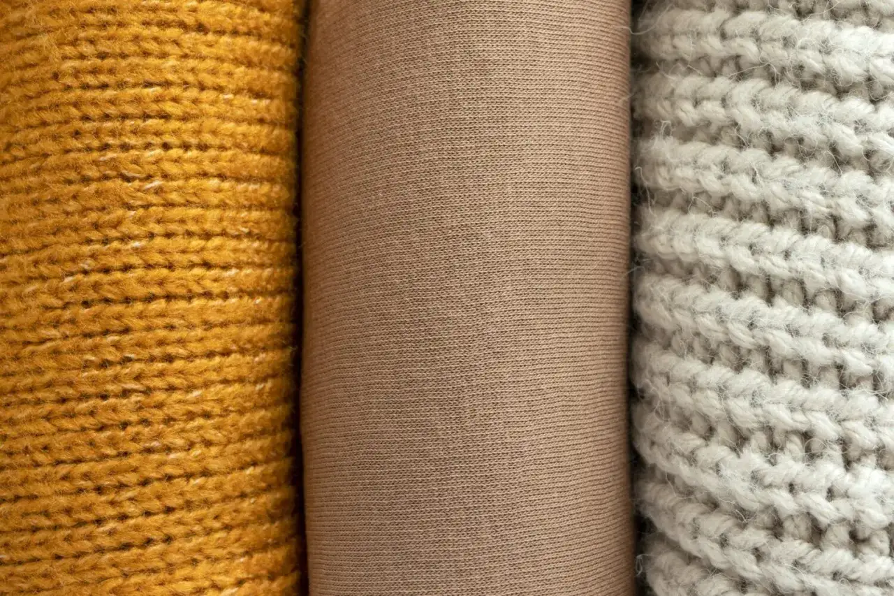 Drei Stoffrollen: gelb gestrickt, beige glatt und hellgrau grob gestrickt. Das beige Material könnte aus Polyester-Plastik sein.