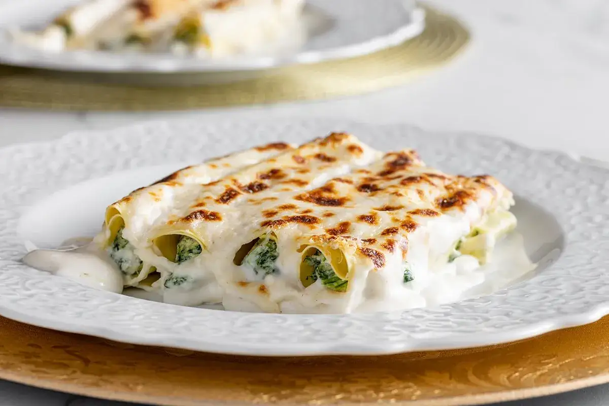 Cannelloni ricotta e spinaci dorati e filanti, serviti su un piatto bianco con una salsa cremosa.