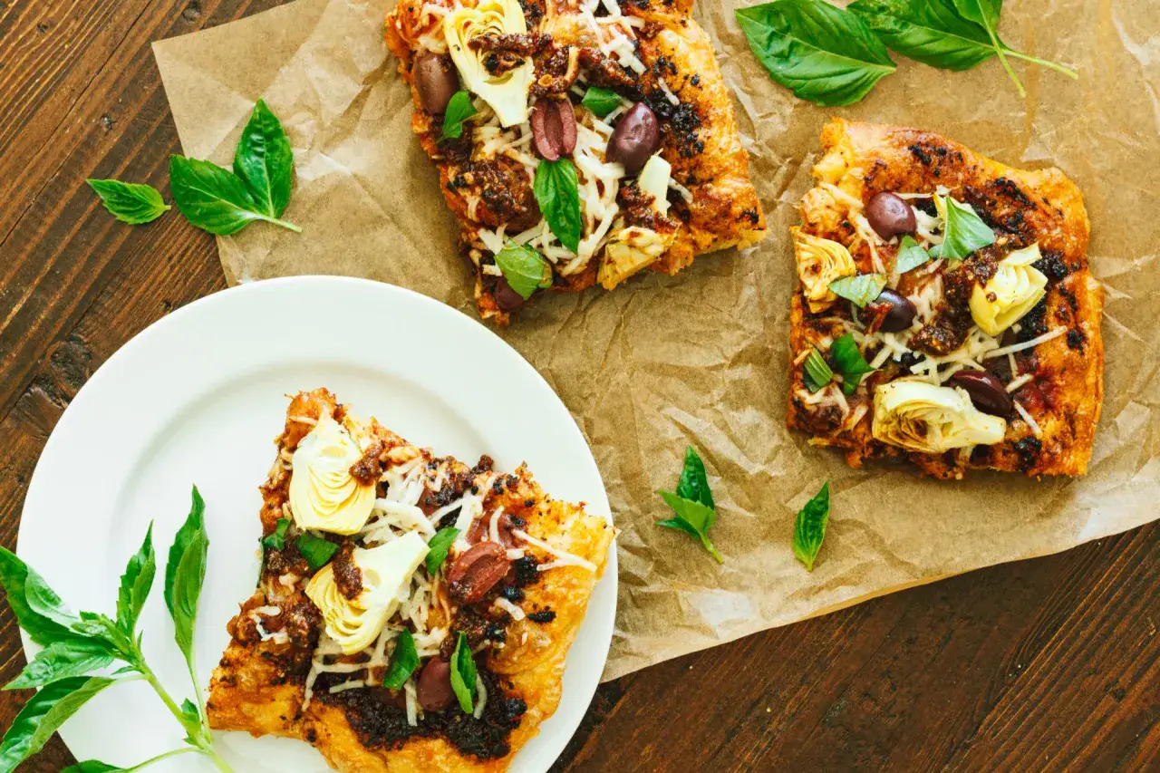 Rebanadas de pizza con alcachofas, aceitunas y queso rallado, con un toque de pesto vegano y hojas de albahaca fresca.