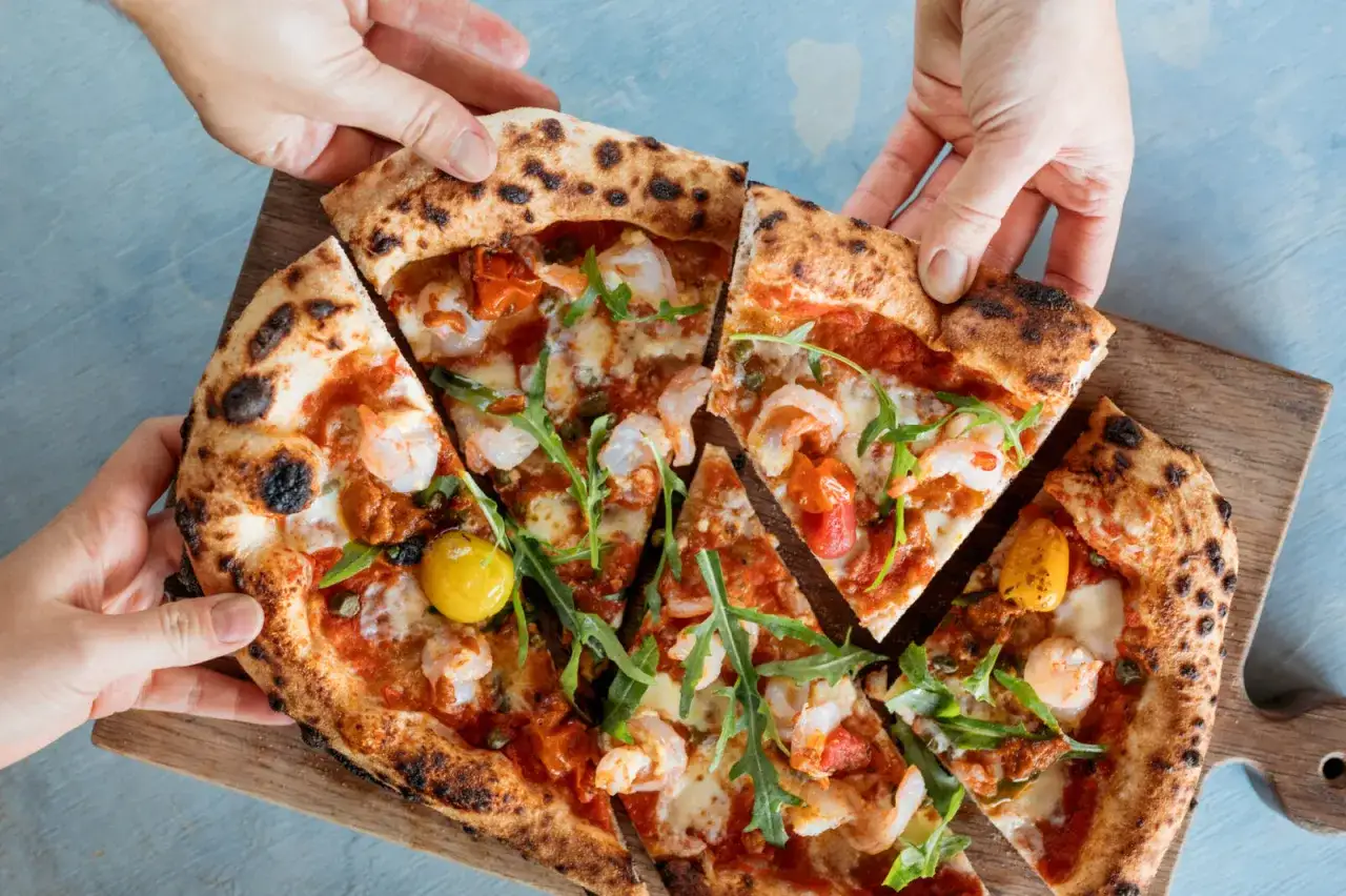 Händer som sträcker sig efter en utsökt italiensk pizza med räkor, tomater och ruccola.
