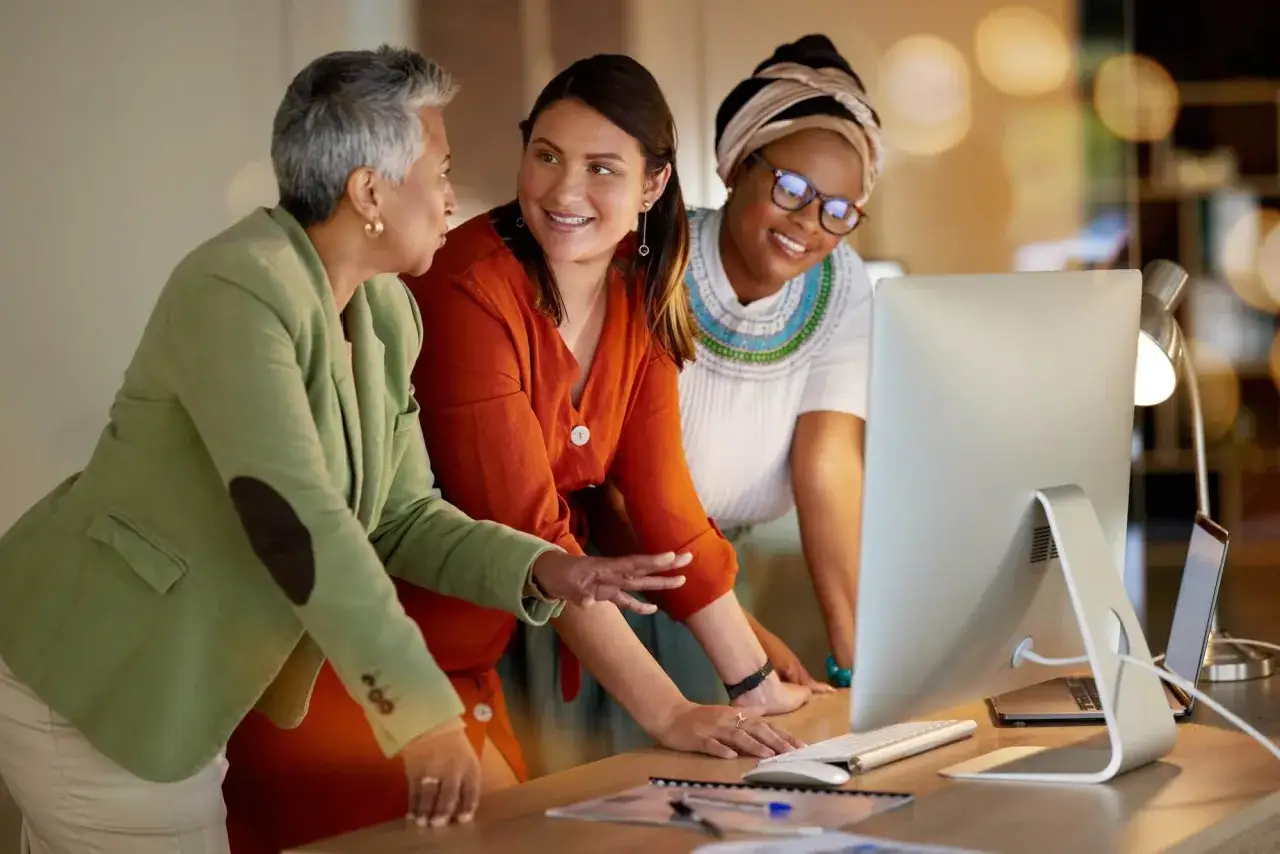 Three women collaborate on a computer, discussing strategies for management and leadership development.