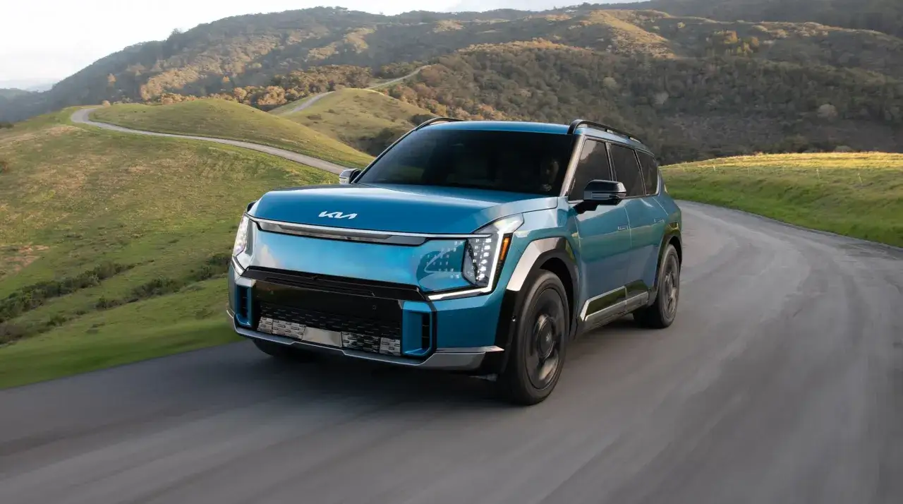 Niebieski Kia EV9 sunie po krętej drodze wśród zielonych wzgórz.