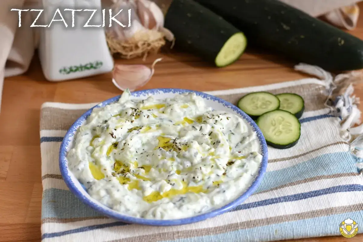 Salsa tzatziki cremosa, condita con olio d'oliva e erbe aromatiche, con cetrioli e aglio sullo sfondo.