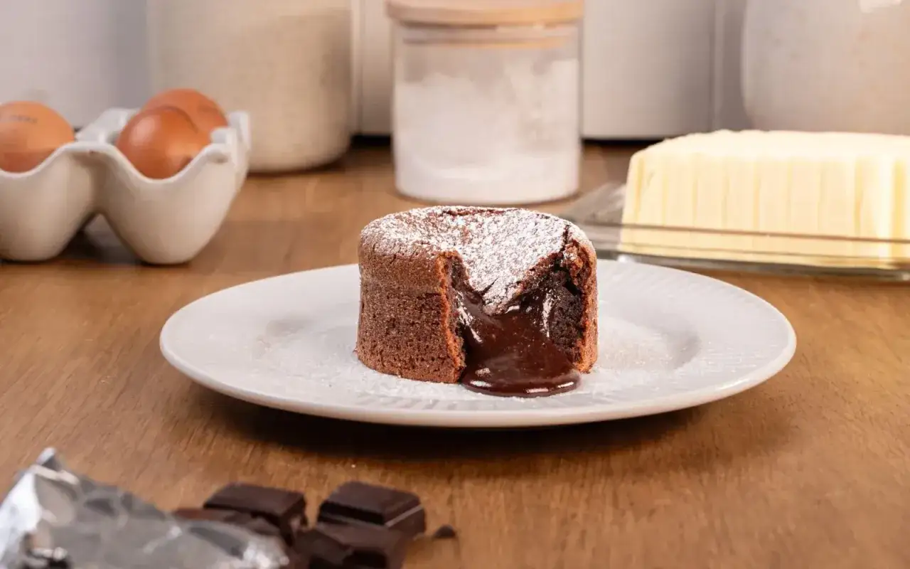 Un délicieux coulant au chocolat, saupoudré de sucre glace, avec son cœur fondant qui coule. Des ingrédients de pâtisserie sont visibles en arrière-plan.
