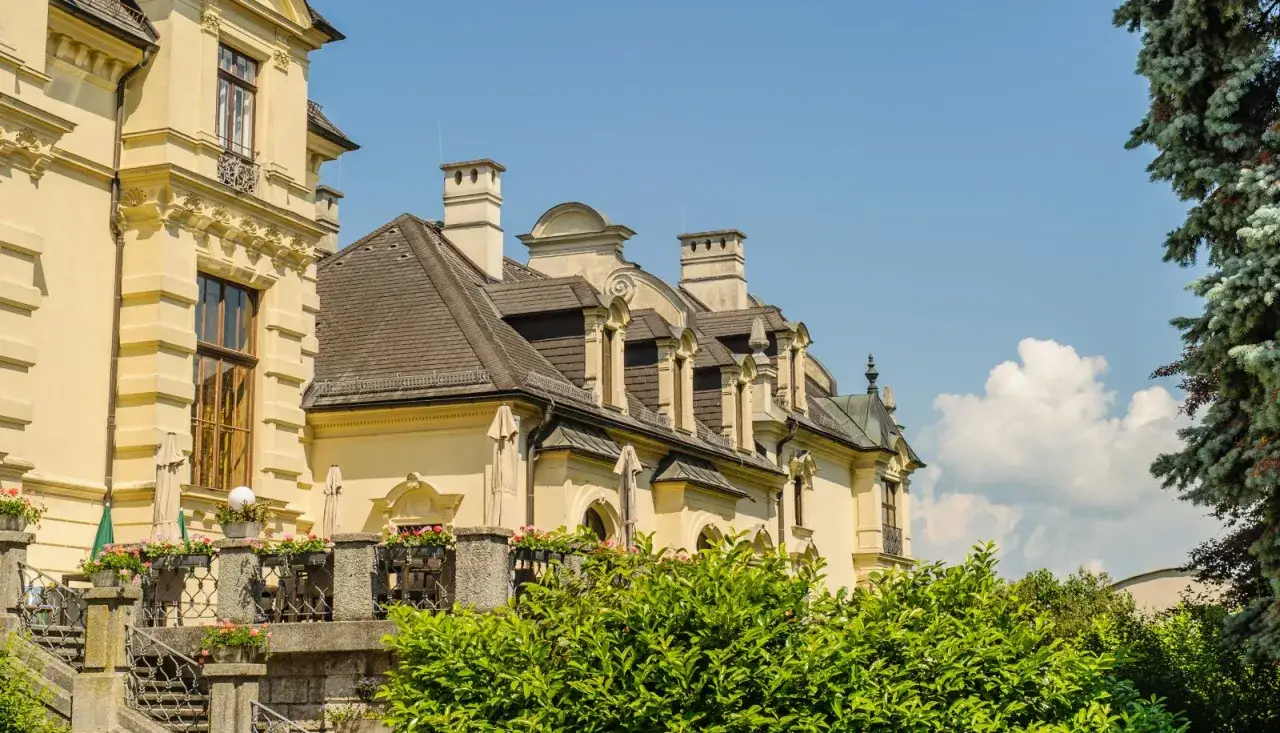 Gelbe Fassade mit detailreicher Architektur und Dachgauben. Ein grüner Baum und blühende Pflanzen umrahmen das Anwesen der Pallottiner Salzburg.